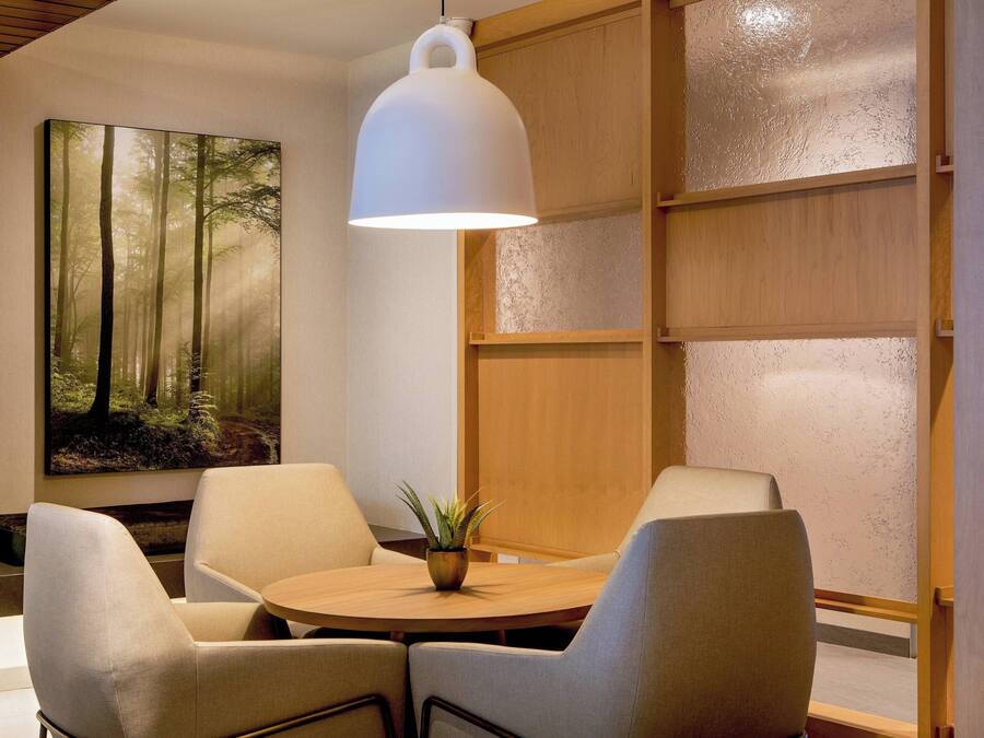 Un interior moderno con una mesa redonda de madera, dos sillas con cojines, una lámpara colgante blanca y un cuadro grande con motivos forestales en la pared.