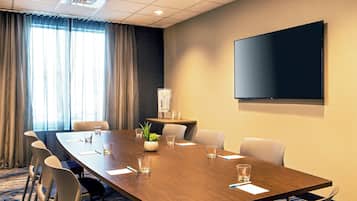 Une salle de conférence avec une longue table, des chaises, un écran de projection et une fenêtre avec des rideaux.