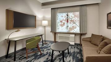 A hotel room with a flat-screen TV mounted on the wall, a desk with a lamp, a green chair, a small round table, and a beige sofa.