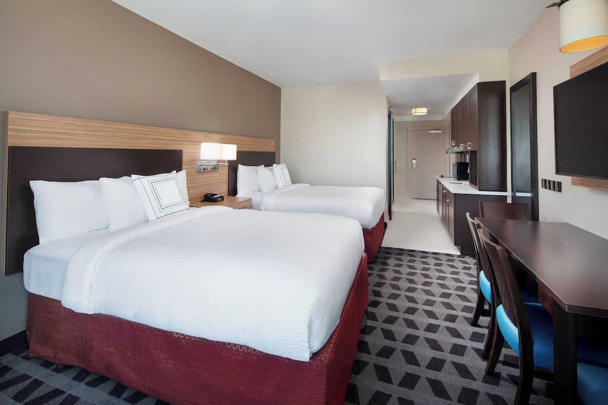 A hotel room with two beds, a desk, a TV, and a kitchenette.