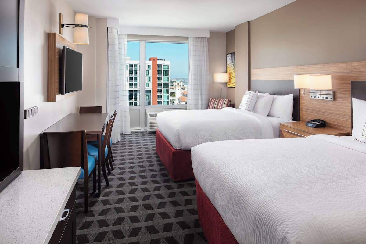 A hotel room with two beds, a desk, a TV, and a window with a city view.