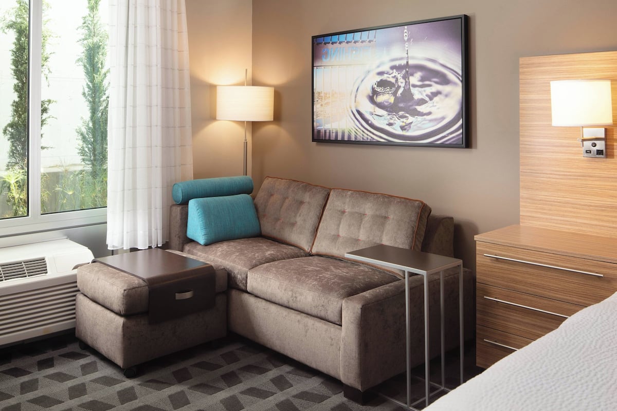 A modern hotel room with a sectional sofa, a small table, a lamp, a painting, and a wooden dresser.