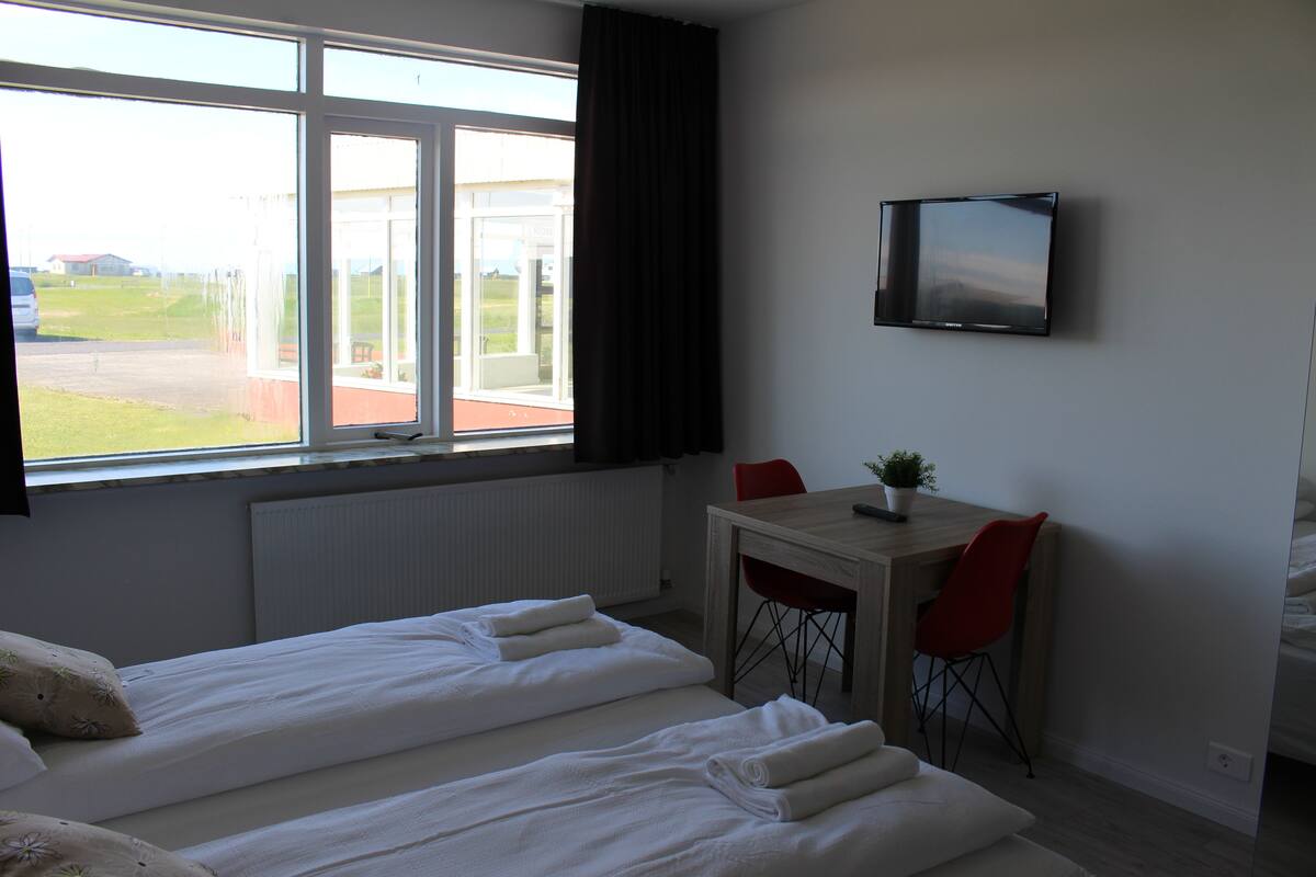 Ein Hotelzimmer mit zwei Betten, einem Fernseher, einem kleinen Tisch mit Stuhl und einem Fenster mit Blick ins Freie.