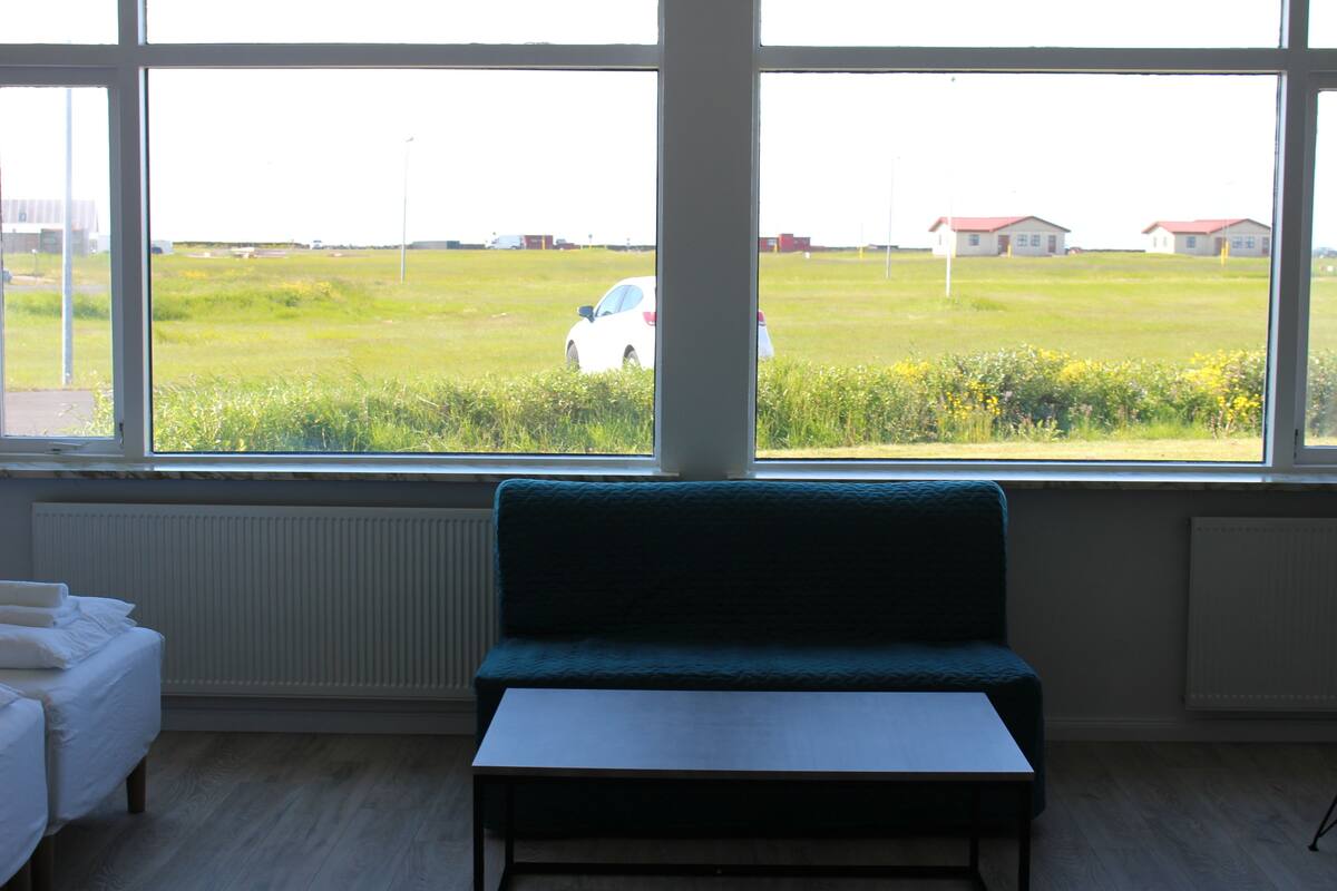 Ein Zimmer mit großem Fenster, einem grünen Sofa, einem weißen Tisch und einem Bett mit weißer Bettwäsche.