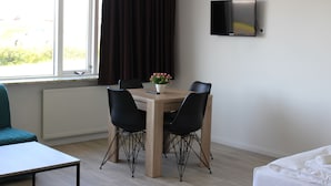 A room with a wooden dining table, four black chairs, a small potted plant, a television on the wall, and a bed with white bedding.