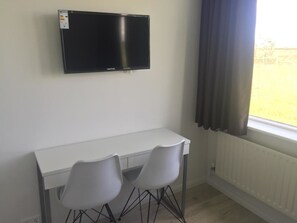A room with a white desk, two white chairs, a flat-screen TV mounted on the wall, and a window with curtains. - Ocean Beach Apartments (Stokkseyri)