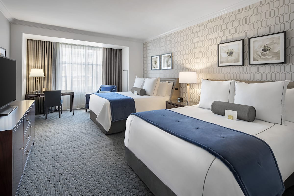 A hotel room with two beds, a desk, a chair, a TV, and artwork on the wall.