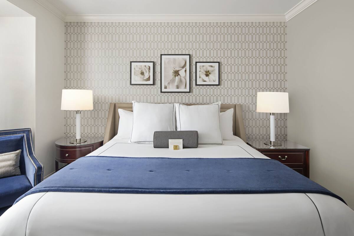 A neatly arranged hotel room with a large bed, two bedside tables with lamps, a blue throw blanket, and a wall with a geometric pattern.