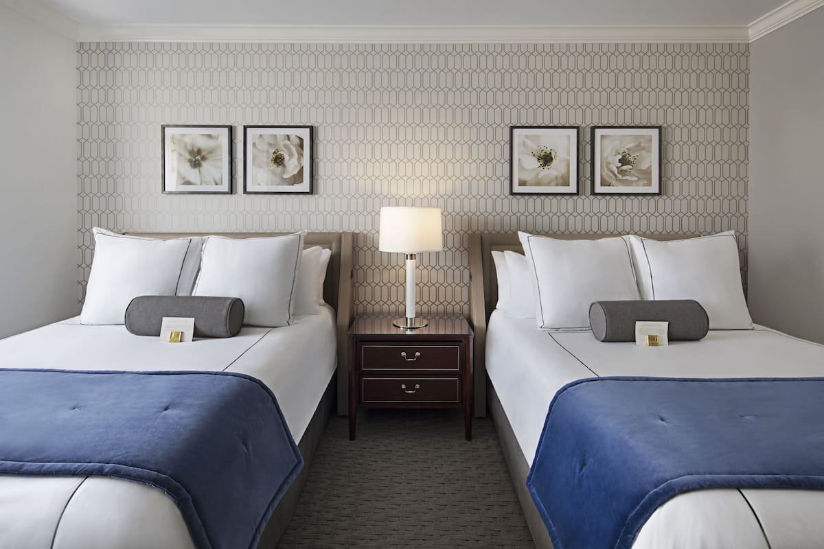 A hotel room with two beds, a nightstand with a lamp, and three framed floral artworks on the wall.
