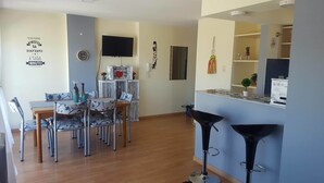 A dining area with a wooden table and chairs, a TV mounted on the wall, and a kitchen area with a counter and stools.