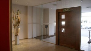 A reception area with a wooden door labeled "SALIDA DE EMERGENCIA" (emergency exit), a glass partition, and a potted plant.