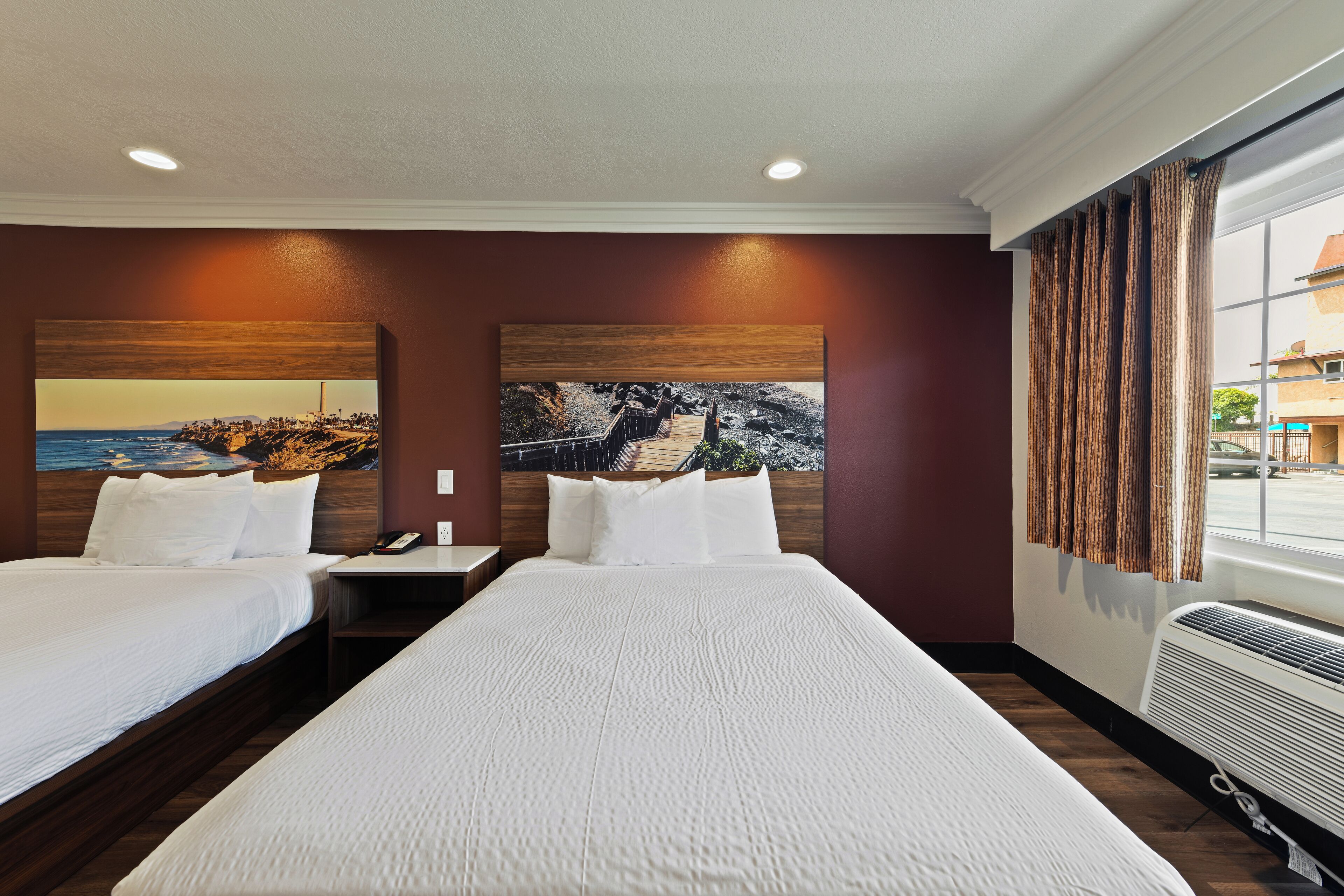 Quarto de hotel com duas camas, um quadro grande na parede, uma mesa de cabeceira e uma janela com cortinas.
