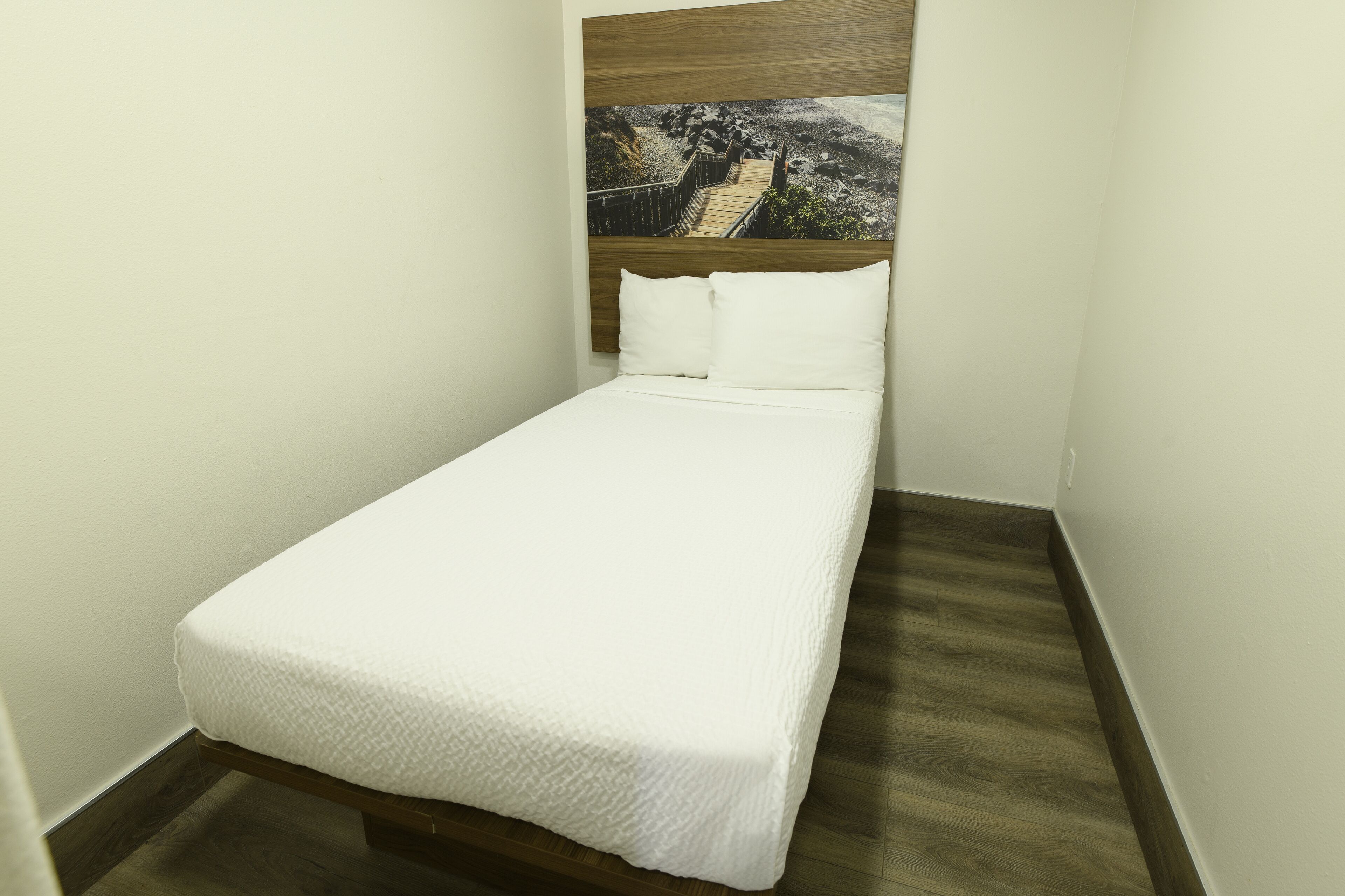 A small, neatly made bed with a white bedspread in a corner room with a wooden floor and a painting above the bed.