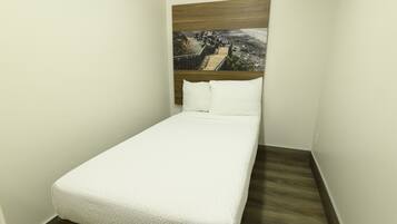 Un letto piccolo e ben rifatto con copriletto bianco in una stanza angolare con pavimento in legno e un quadro sopra il letto.