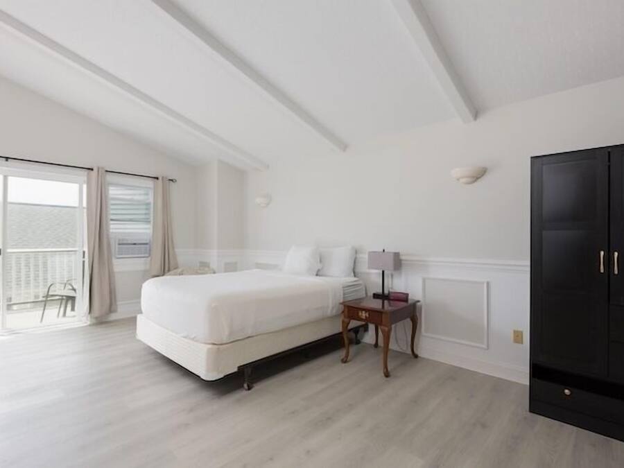 A bedroom with a bed, a nightstand, a lamp, a wardrobe, and a sliding glass door leading to an outdoor area.