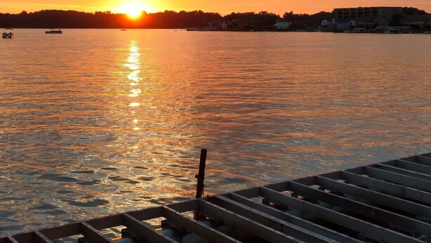 A sunset over a calm body of water with boats, a dock, and a distant shoreline.