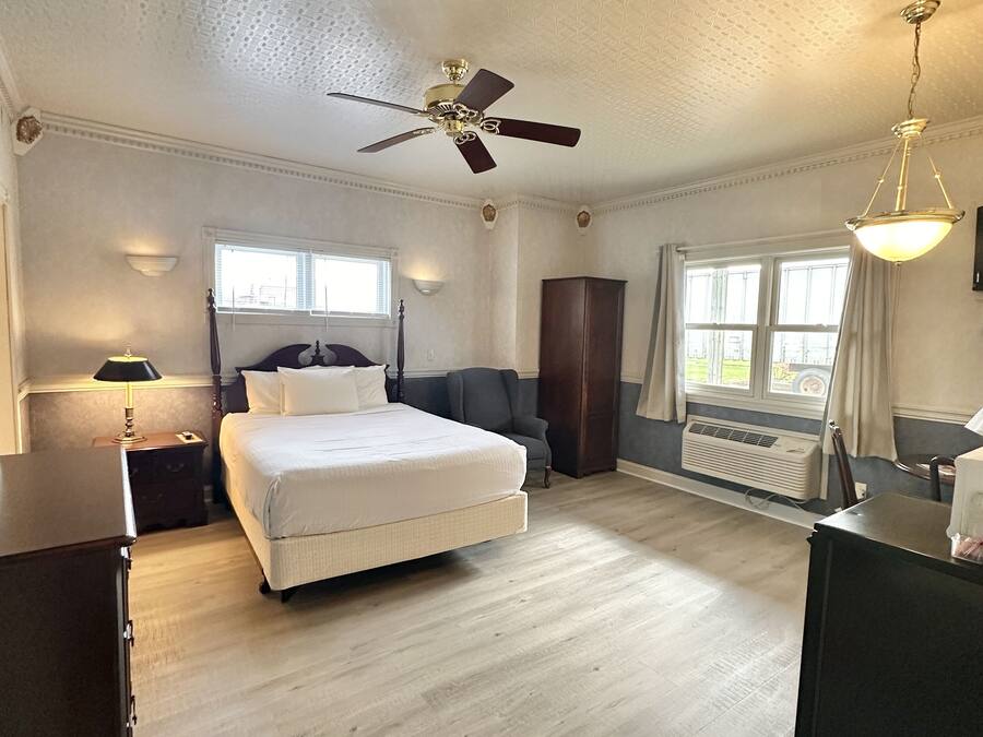 Une chambre spacieuse avec un grand lit, un ventilateur de plafond, une fenêtre avec des rideaux et un radiateur.