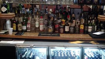 Uma área de bar com prateleiras de garrafas de bebidas e copos, um balcão de atendimento e um refrigerador com diversas bebidas.