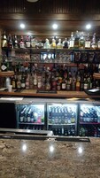 Una zona de bar con estantes llenos de botellas de licor y vasos, una barra de servicio y un enfriador con diversas bebidas.