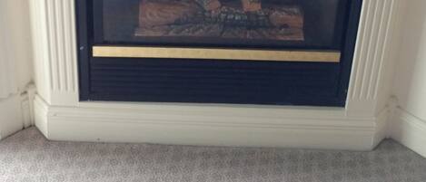 A corner fireplace with a white mantel and a glass door, displaying a fire.