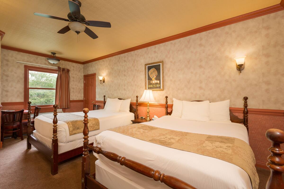 A hotel room with two wooden beds, a ceiling fan, a window with curtains, and a wall-mounted lamp.