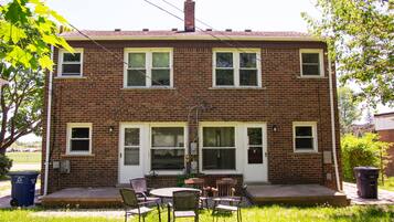 A two-story brick house with a front porch, a small patio with outdoor furniture, and a visible trash can.