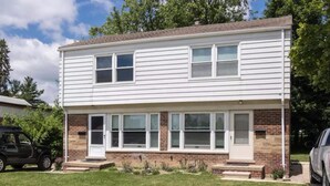 A two-story house with a brick facade and white siding, featuring a front porch with steps and a small garden area.