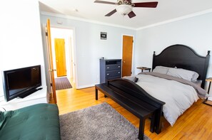 Een slaapkamer met een bed, een televisie, een plafondventilator en een deur naar een andere kamer.