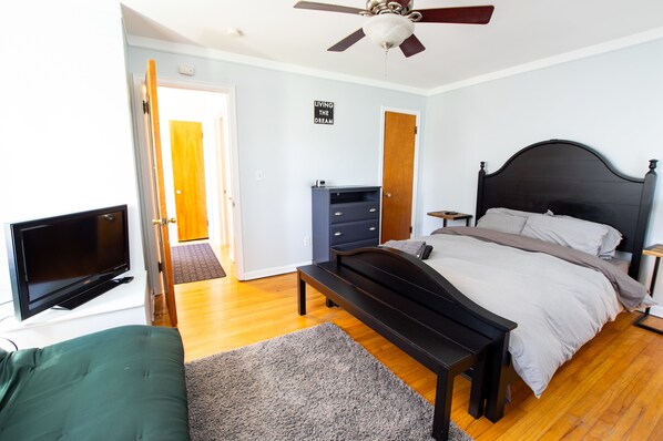 A bedroom with a bed, a TV, a ceiling fan, and a door leading to another room.