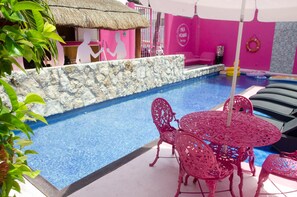 Un área de piscina con un muro de piedra, una mesa con sillas y una estructura cubierta de paja.