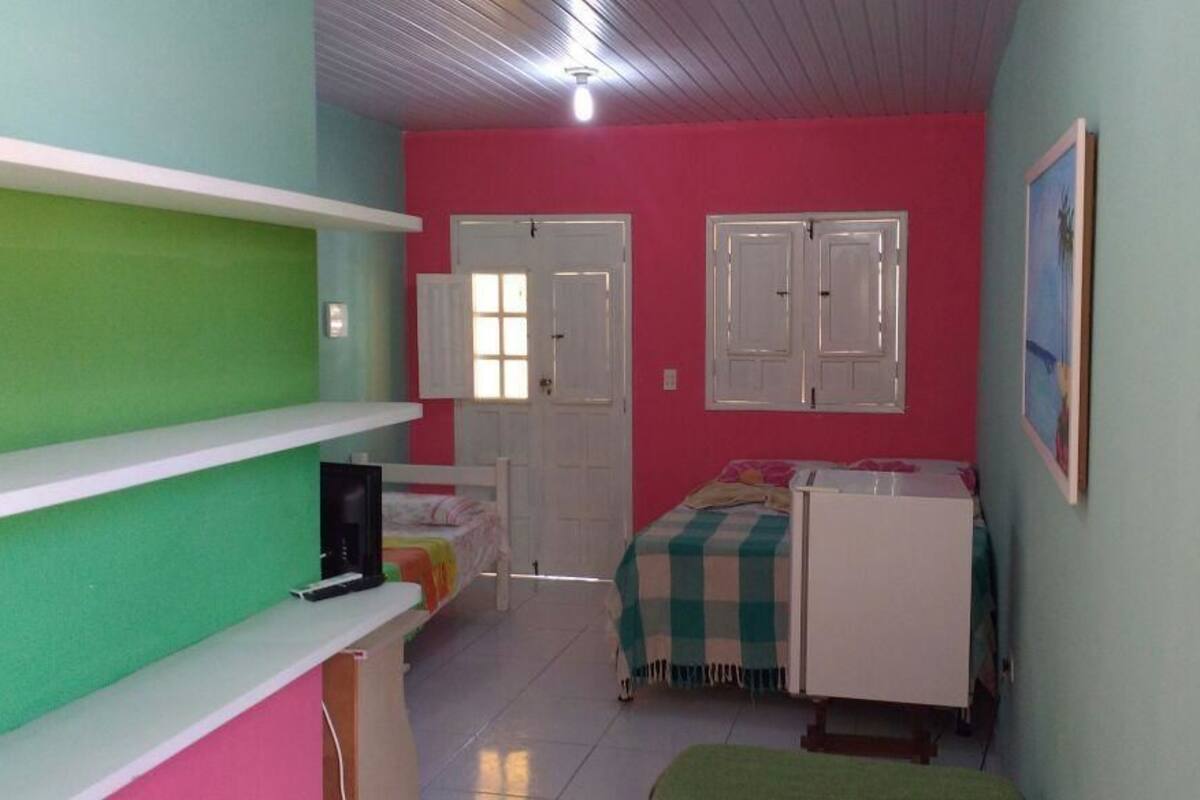 Una habitació petita i colorida amb un llit, un escriptori i una cadira. Hi ha prestatges a la paret i una porta amb una finestra.