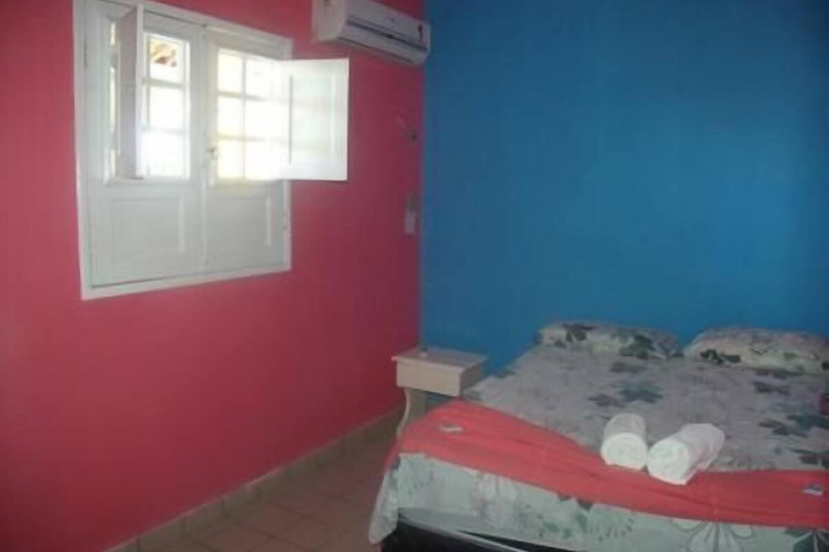 Una habitació amb un llit, una finestra amb persianes blanques i un aparell d'aire condicionat fixat a la paret.