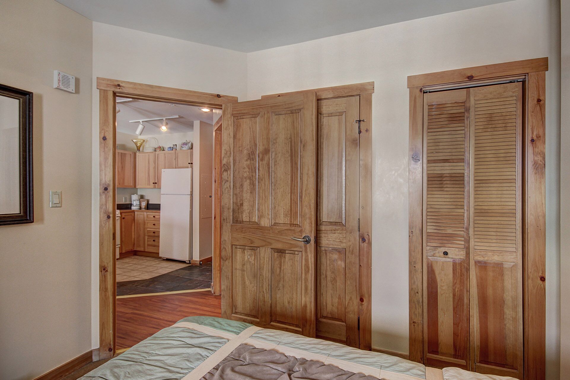 Une pièce comprenant un lit, une armoire en bois et un coin cuisine visible à travers une porte ouverte.