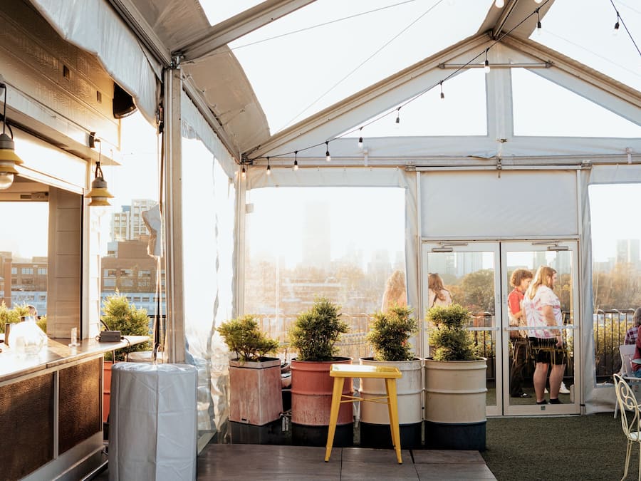 An outdoor dining area with a high ceiling and string lights, surrounded by potted plants and a city view.