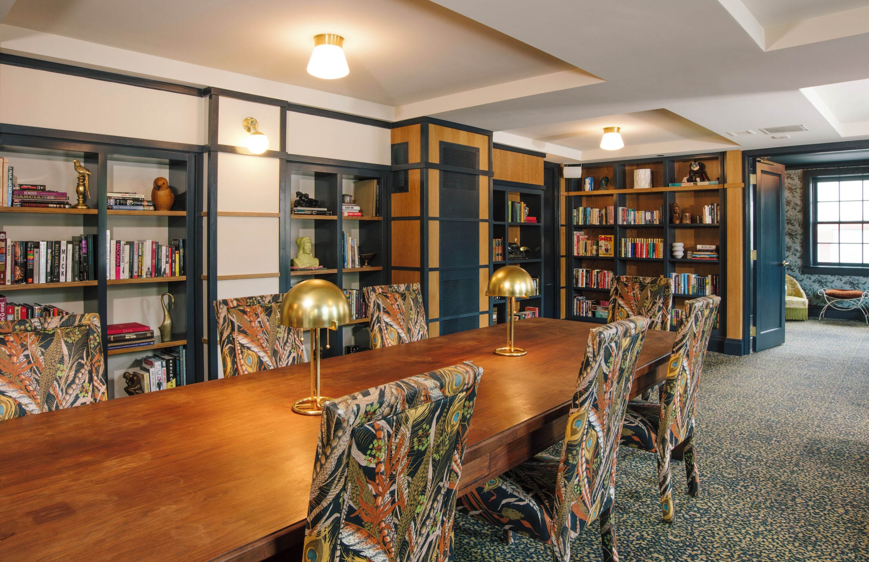 Eine Bibliothek mit einem langen Holztisch und gemusterten Stühlen, umgeben von Bücherregalen voller Bücher und Dekoration.