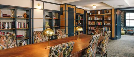 A library with a long wooden table and patterned chairs, surrounded by bookshelves filled with books and decorative items.