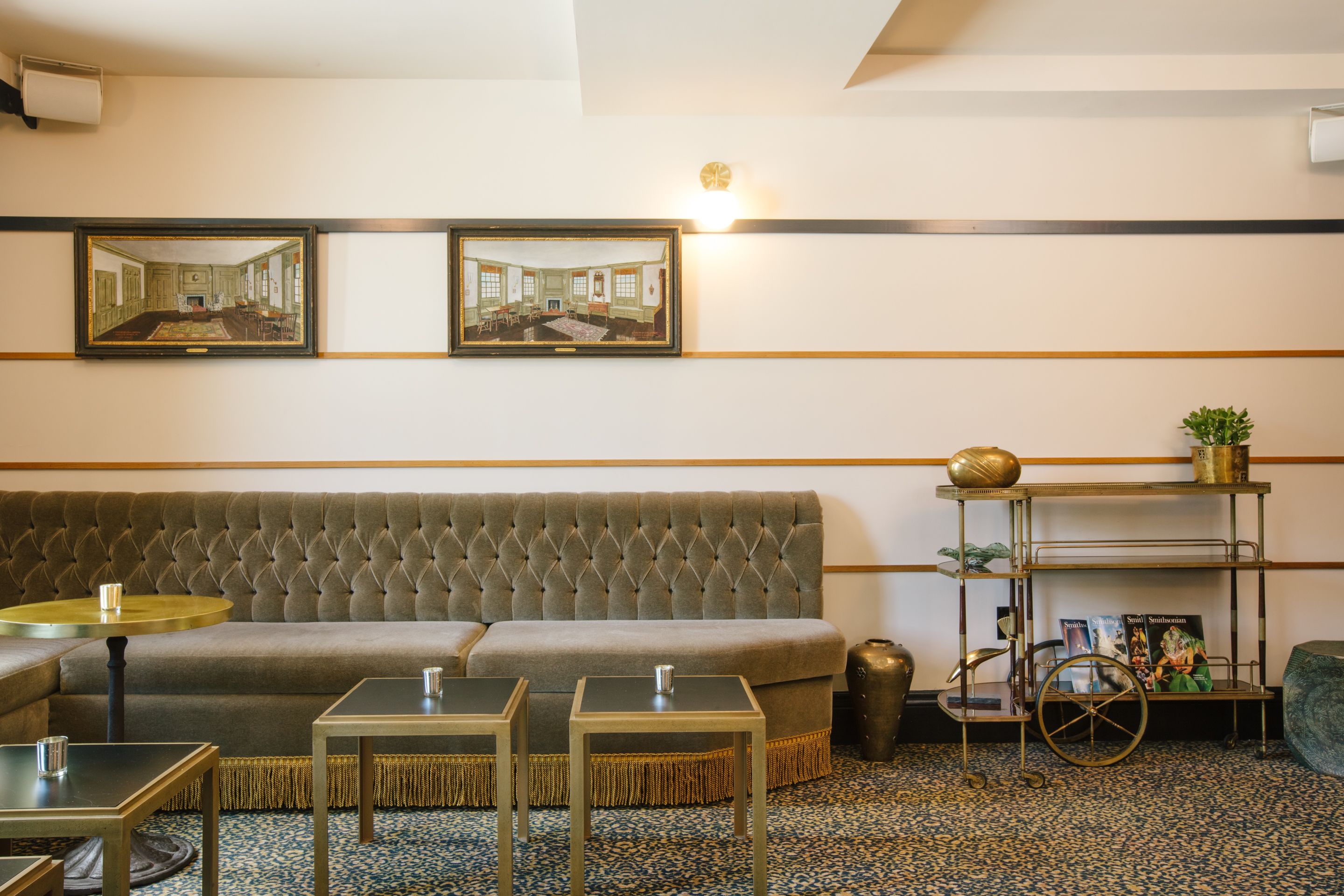 Een kamer met een geborduurde bank, twee kleine tafels, een decoratieve wagen met boeken en lijstjes met afbeeldingen aan de muur.