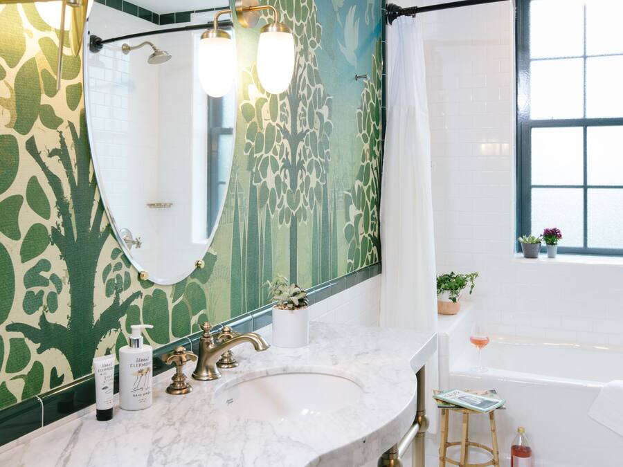A bathroom with a large mirror, a sink with a gold faucet, a white bathtub, and a window with potted plants.
