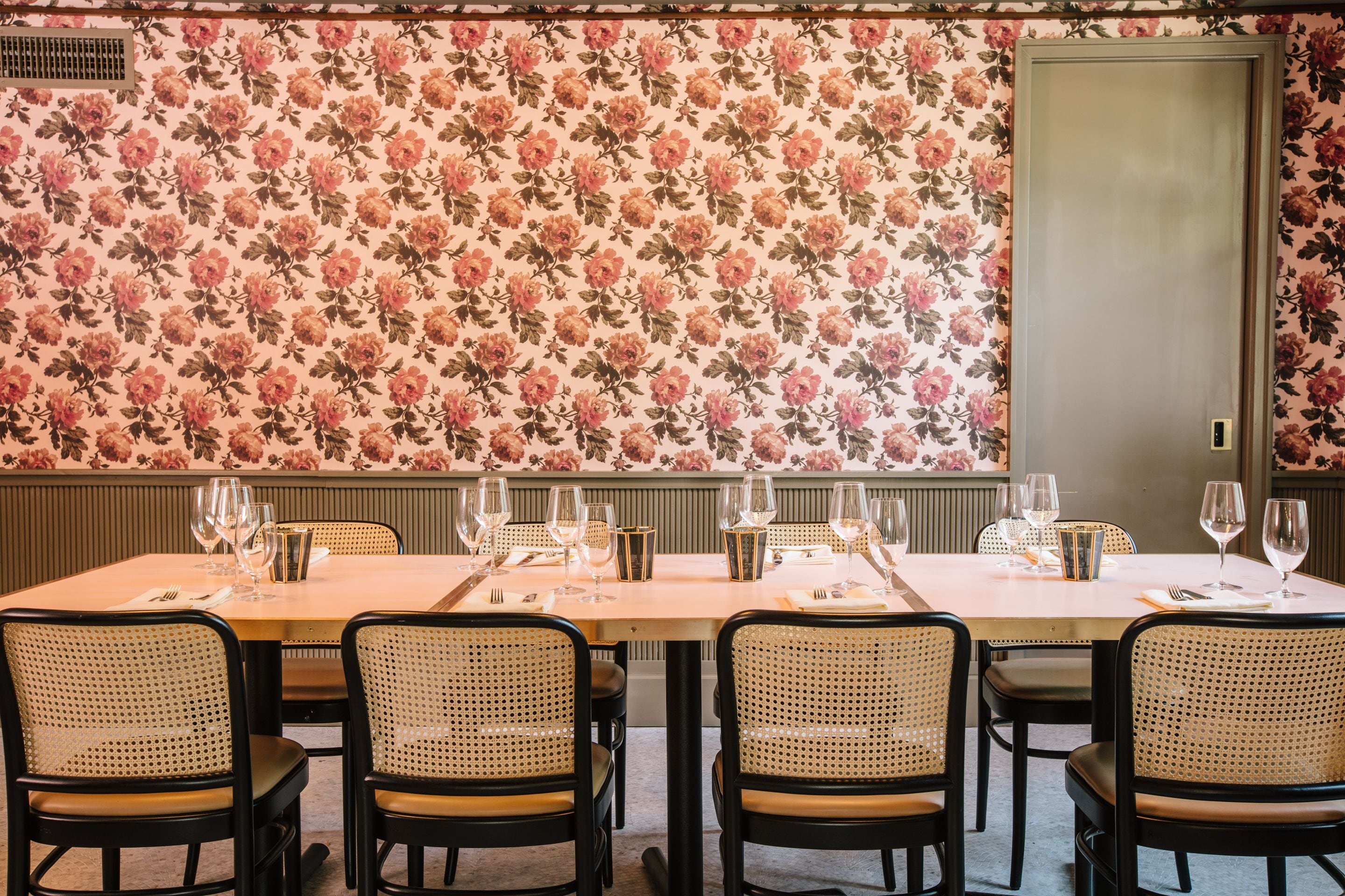 A dining area with a floral wallpaper, a long table set for six, and black and beige chairs.