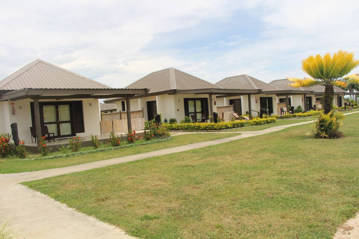 A row of modern, single-story buildings with flat roofs and large windows, surrounded by well-maintained lawns and a variety of plants.