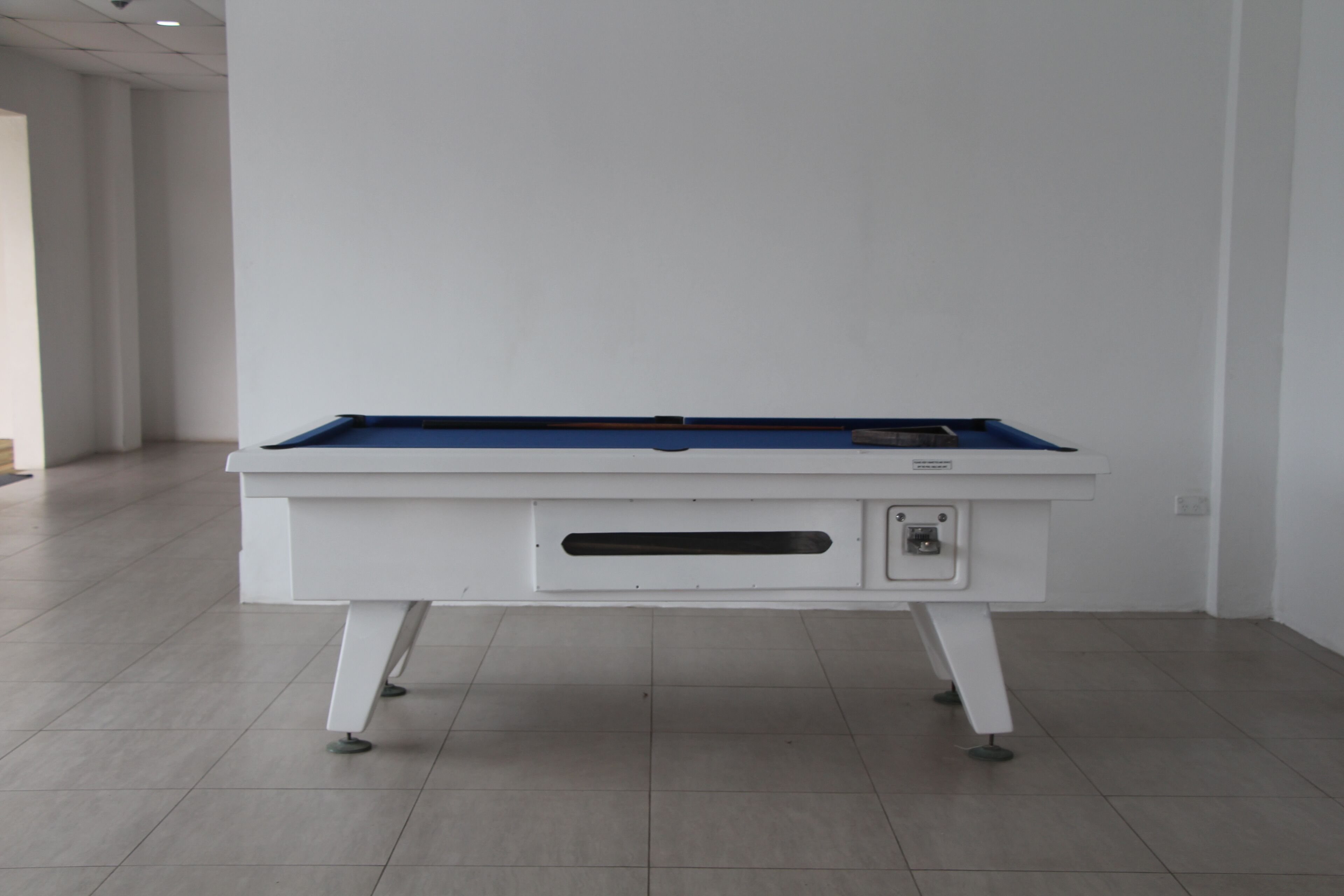 A white pool table with a blue felt top, positioned in a room with white walls and a tiled floor.