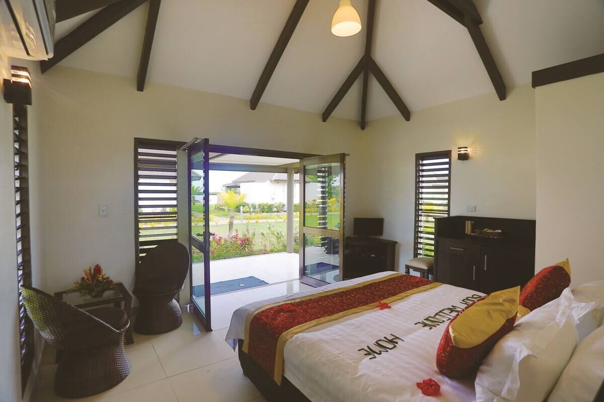 Quarto com cama, escrivaninha, cadeira e vista para um jardim através de uma porta de vidro.