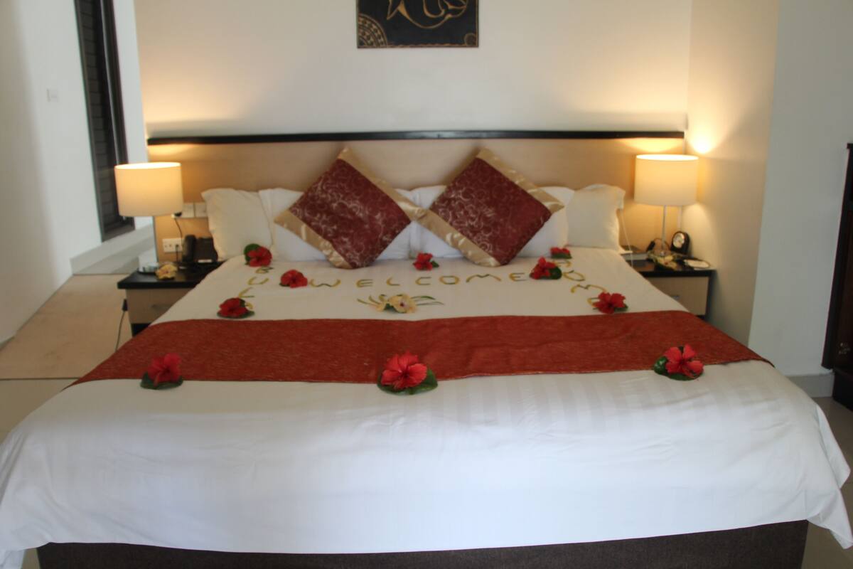 A neatly made bed with a 'WELCOME' sign, red and beige pillows, and red flowers on the bedspread.