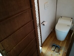 A small bathroom with a toilet, a paper towel dispenser, and a pair of slippers on the floor. - Guest House Kamejikan (Kamakura)
