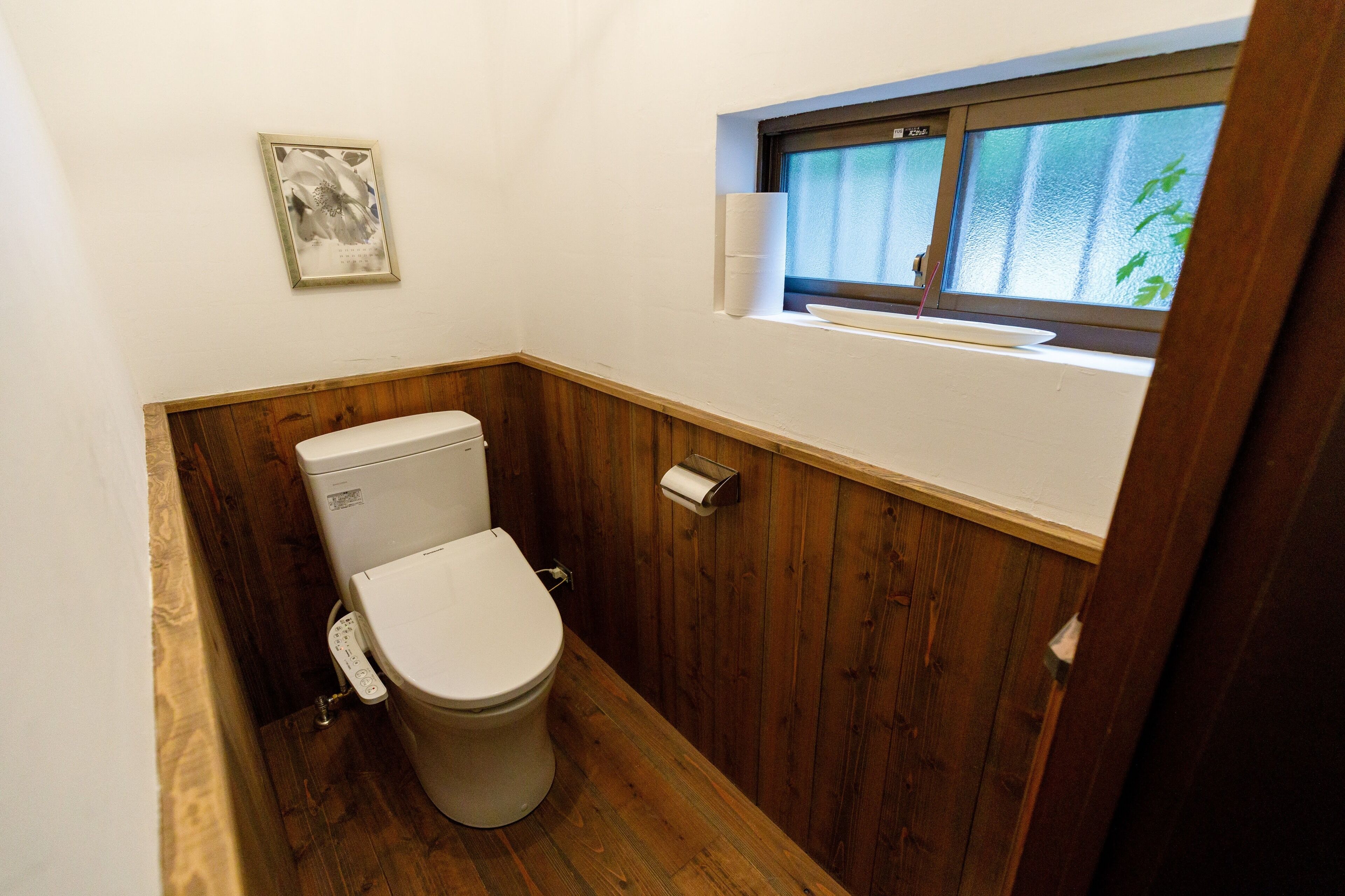 Ein kleines Badezimmer mit Toilette, Holzvertäfelung, einem Fenster mit Blick und einem gerahmten Bild an der Wand.