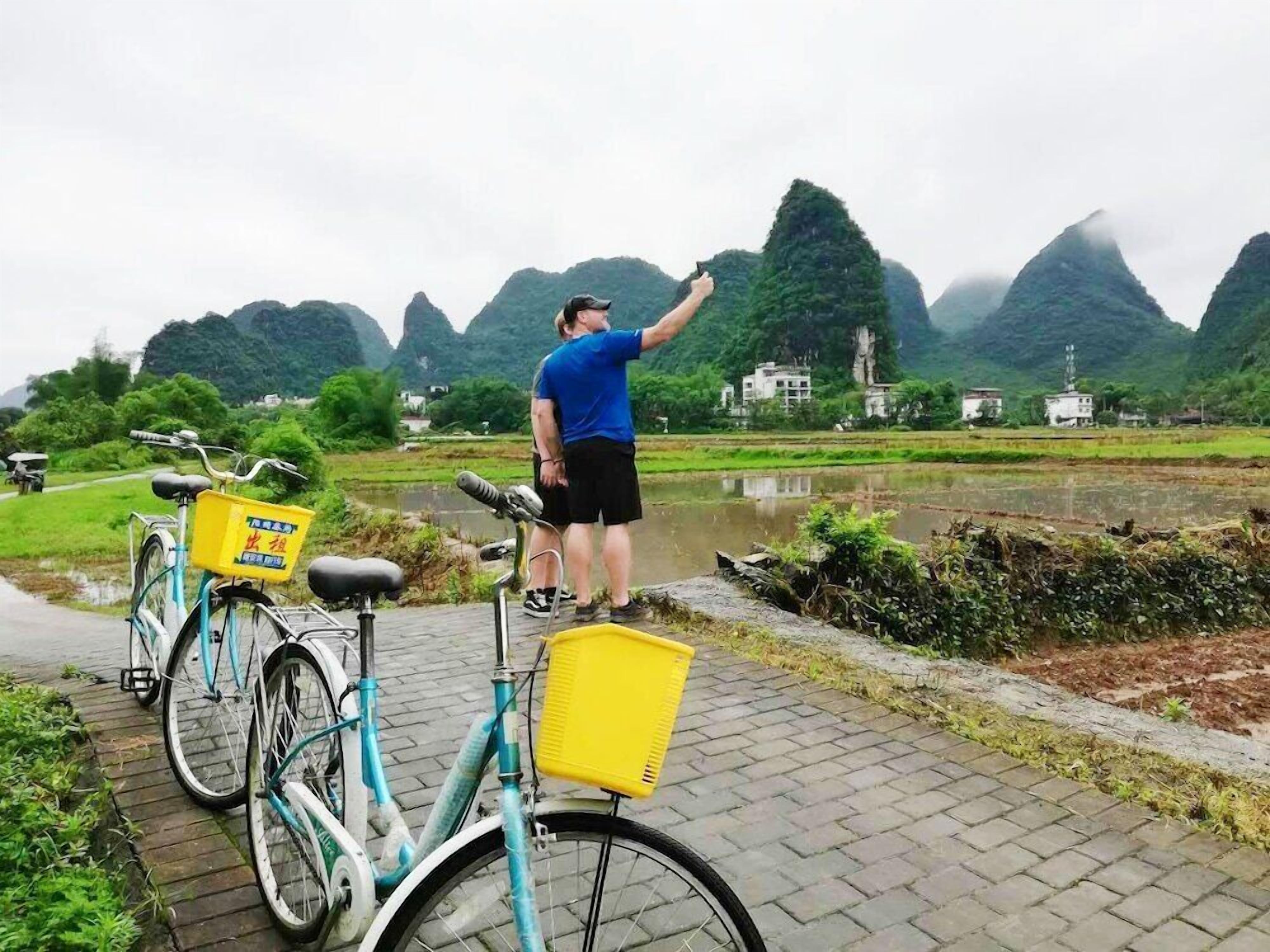 A person standing by a bicycle with a yellow basket, overlooking a rural landscape with fields and mountains.
