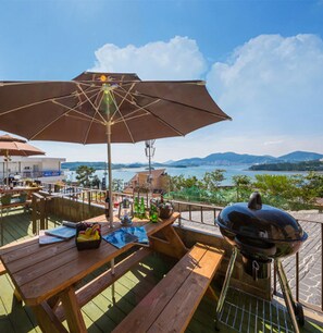 An outdoor dining area with a wooden table, a barbecue grill, and an umbrella providing shade. - Yeosu Ggotnal Pension (Yeosu)