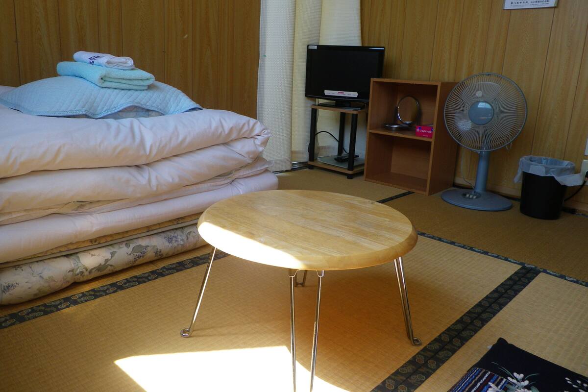 A hotel room with a bed, a small table, a TV, a fan, and a trash bin.