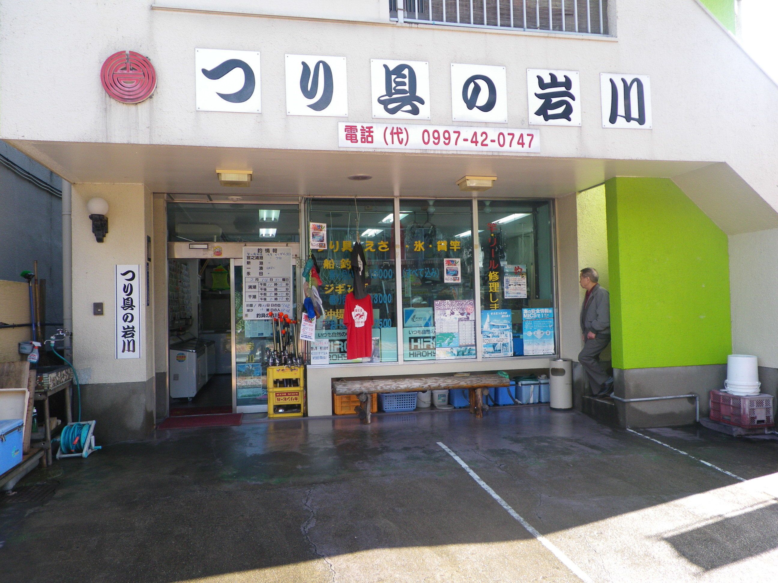 A storefront with Japanese signage, a contact phone number, and various items displayed outside.