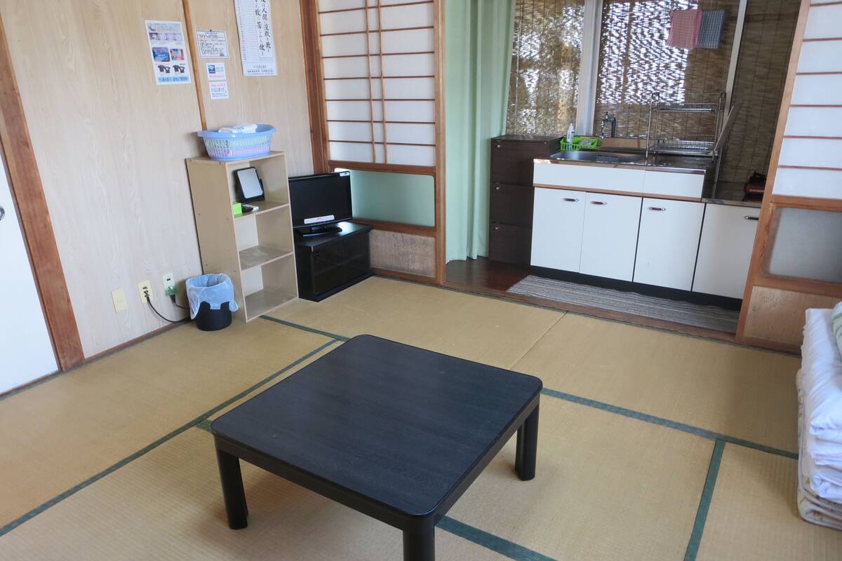 A traditional Japanese room with tatami flooring, a low table, a small TV on a shelf, and a kitchen area with a sink and cabinets.